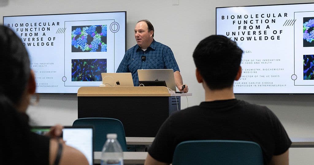 Dr. Justin Siegel in front of an audience with two screens behind him showing the title of his talk, Biomolecular Function from a Universe of Knowledge